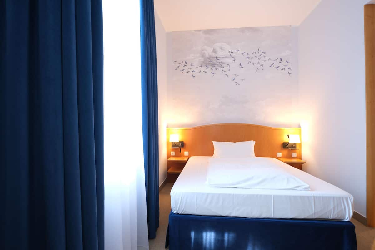 A hotel room with a neatly made bed, two bedside tables with lamps, a wall mural, and a window with curtains.