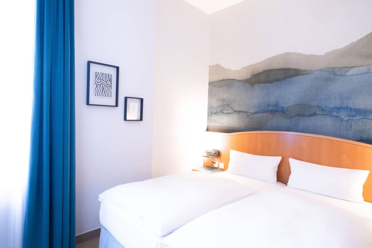 A hotel room with a large bed, a nightstand, a wall-mounted artwork, and a blue curtain.