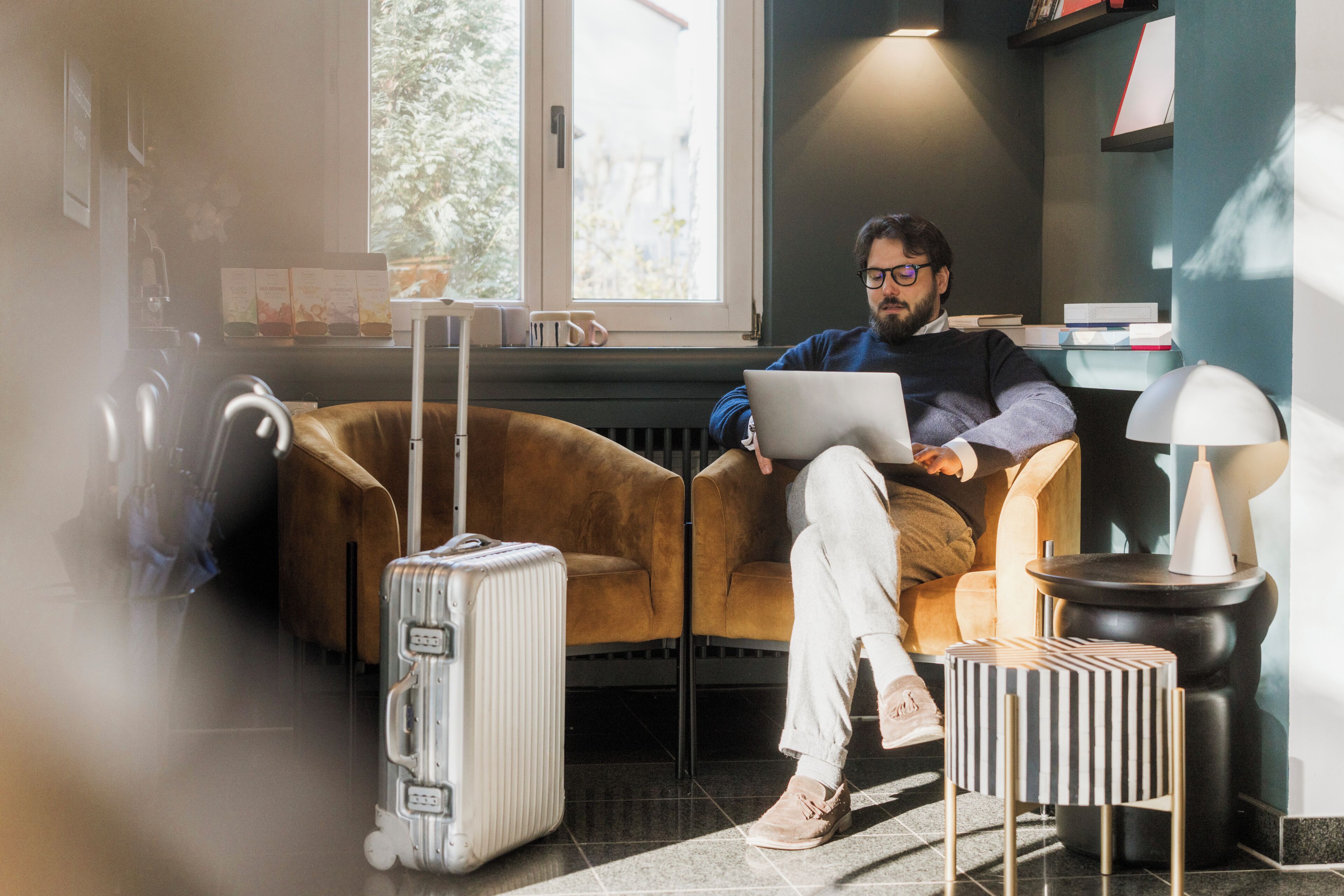 Eine Person sitzt auf einem Sofa mit einem Laptop, einem Koffer und einer Lampe auf einem Tisch.