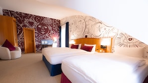 A hotel room with two beds, a desk, and a chair. The room has patterned wallpaper and a large window. - Hotel Das Lola (Essen)