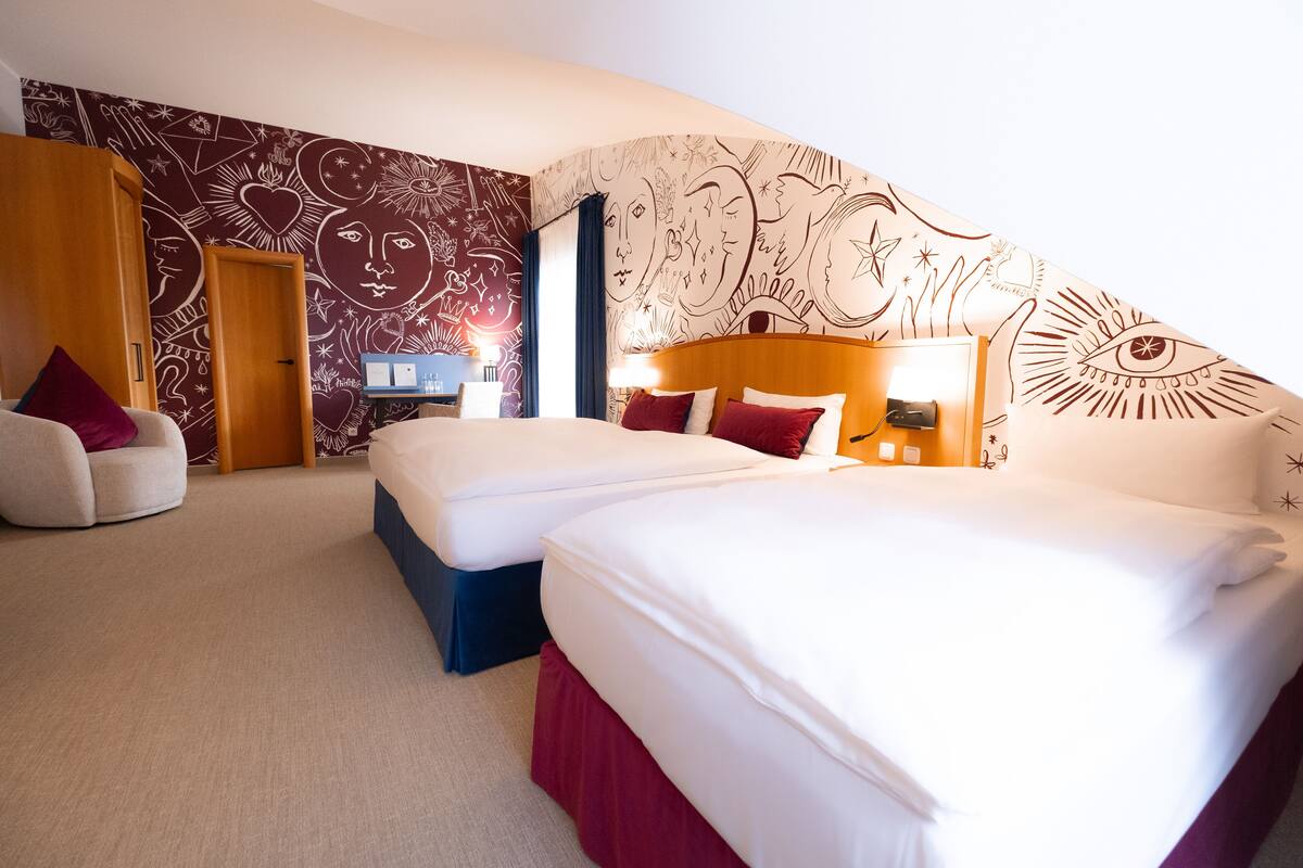 A hotel room with two beds, a desk, and a chair. The room has patterned wallpaper and a large window.