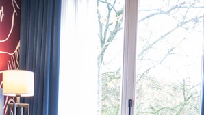A room with a patterned armchair, a table with a lamp, and a view of trees through a glass door. - Hotel Das Lola (Essen)