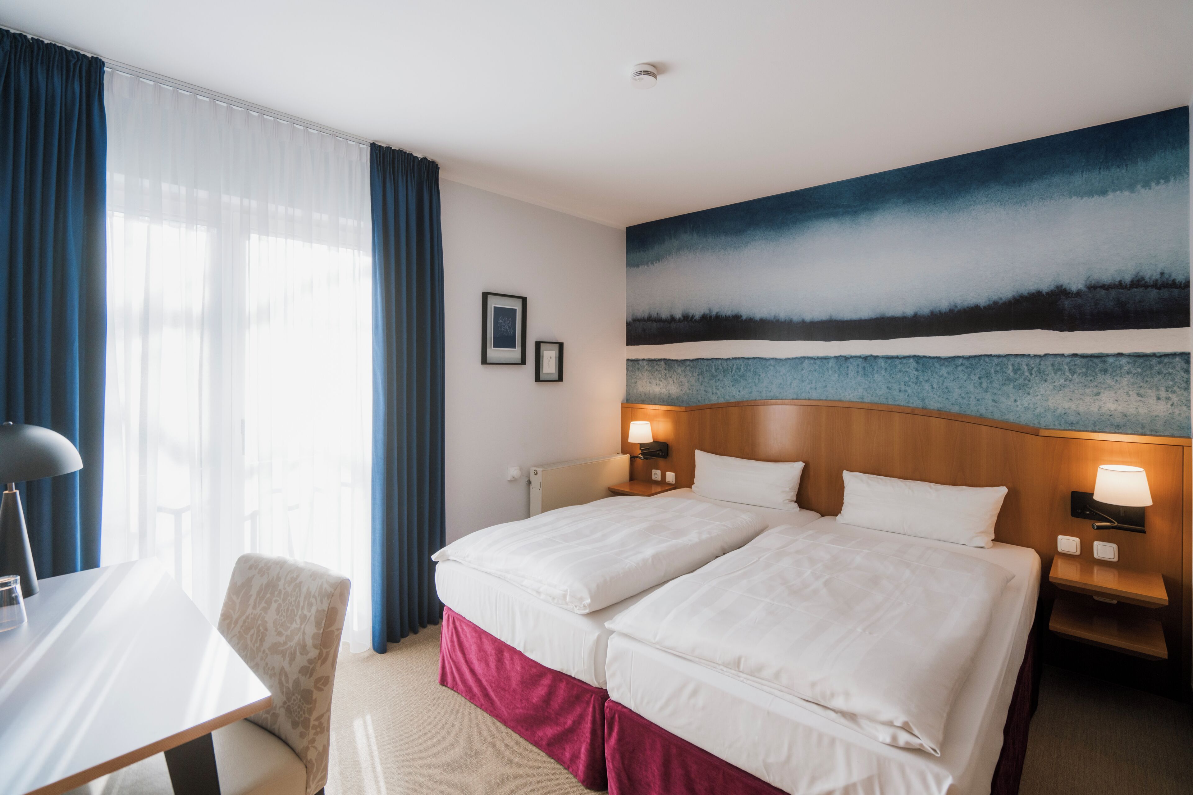 A hotel room with a double bed, a desk, a chair, a nightstand with a lamp, and a painting on the wall.