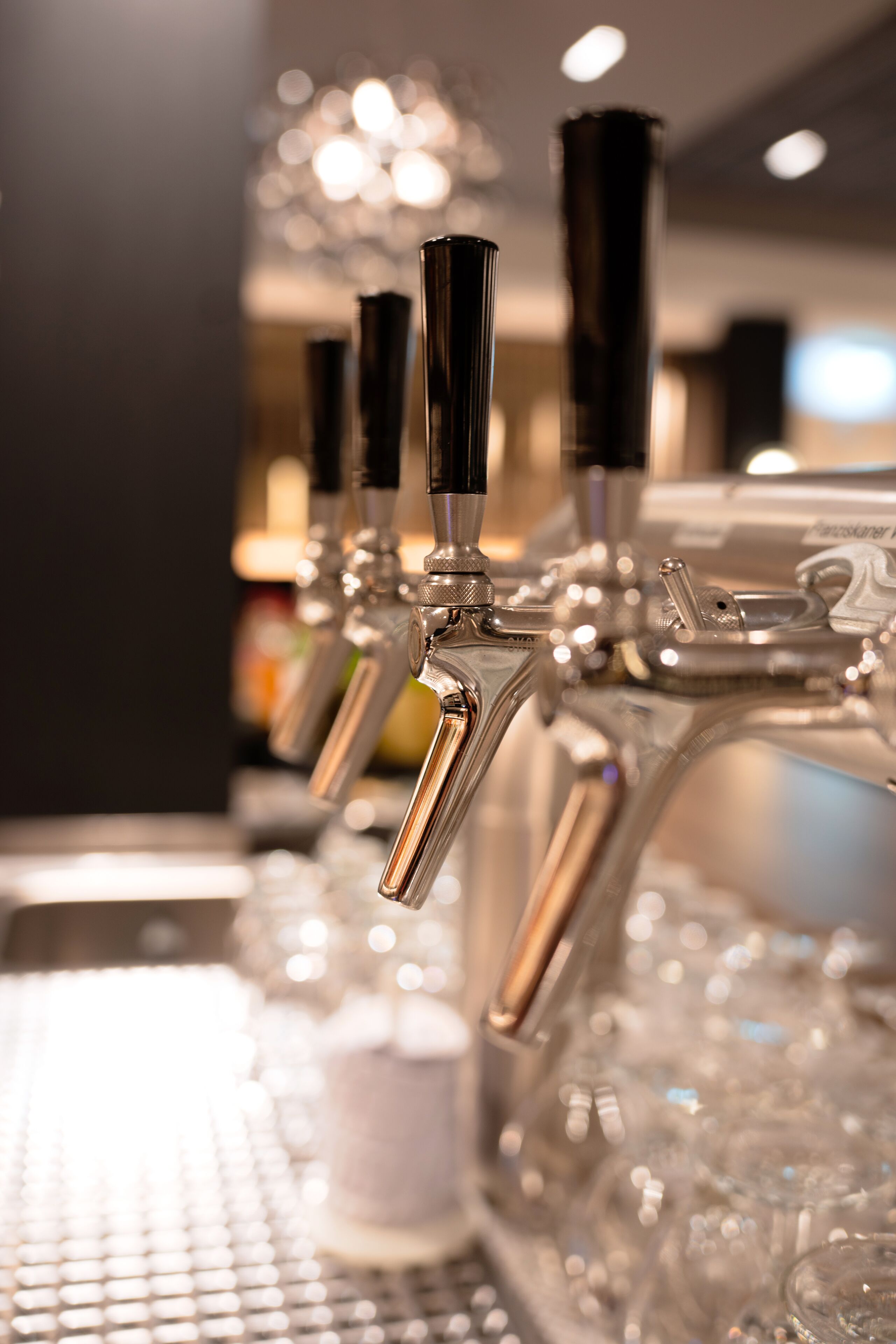 Een rij biertaps met een metalen afwerking en zwarte handgrepen, voor een onscherpe achtergrond met een zachte lichtbron.