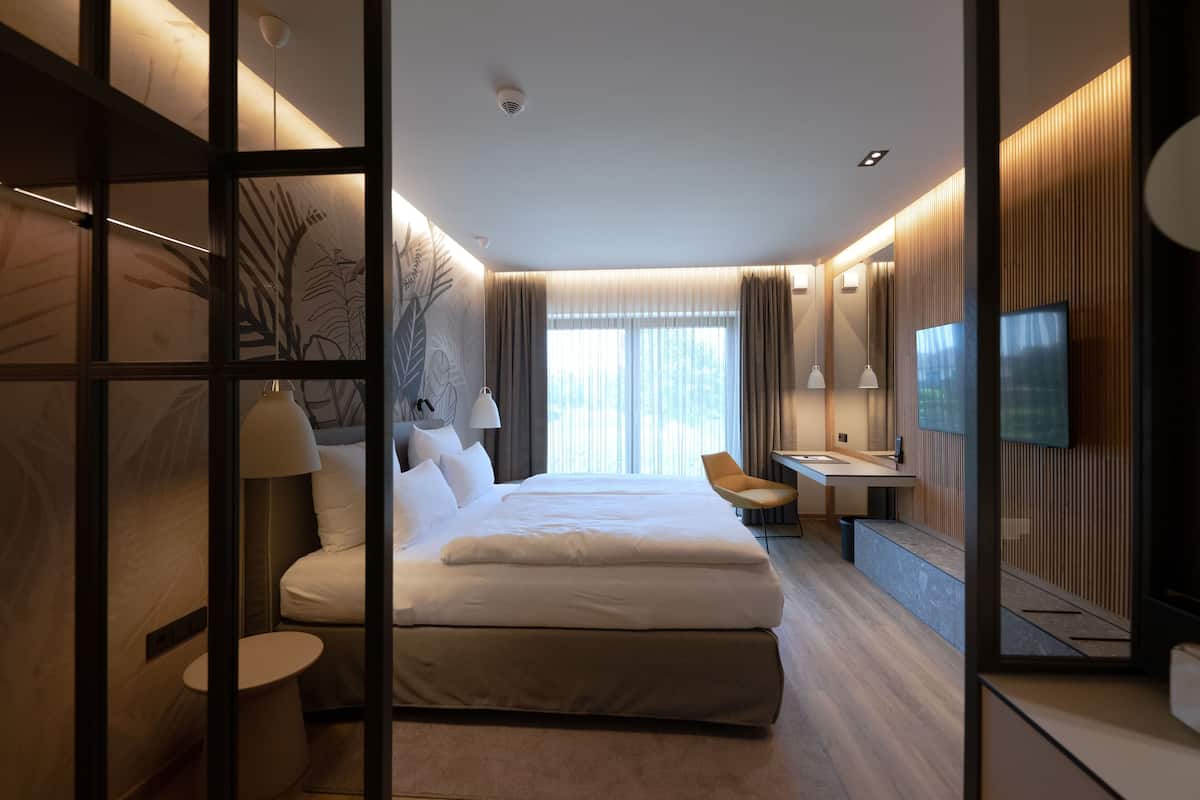 A modern hotel room with a large bed, a sitting area with a desk and chair, a television, and a large window with curtains.