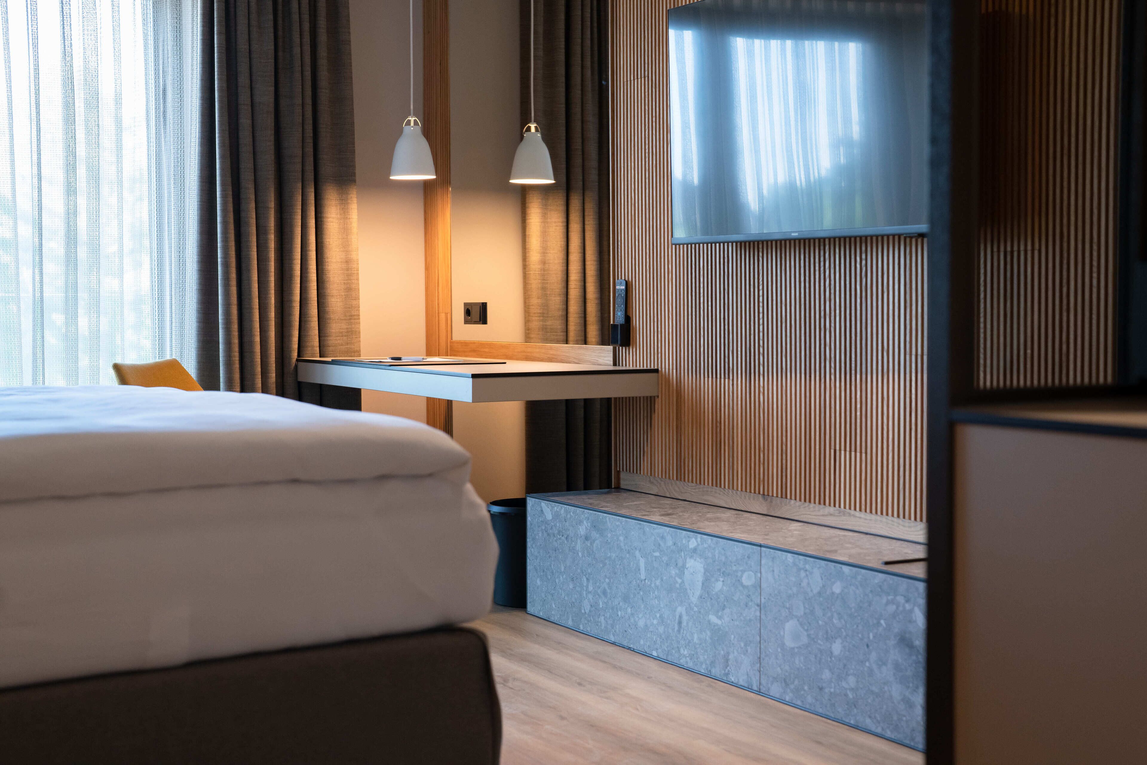 A modern hotel room with a neatly made bed, a desk with a chair, a television mounted on the wall, and a large window with curtains.