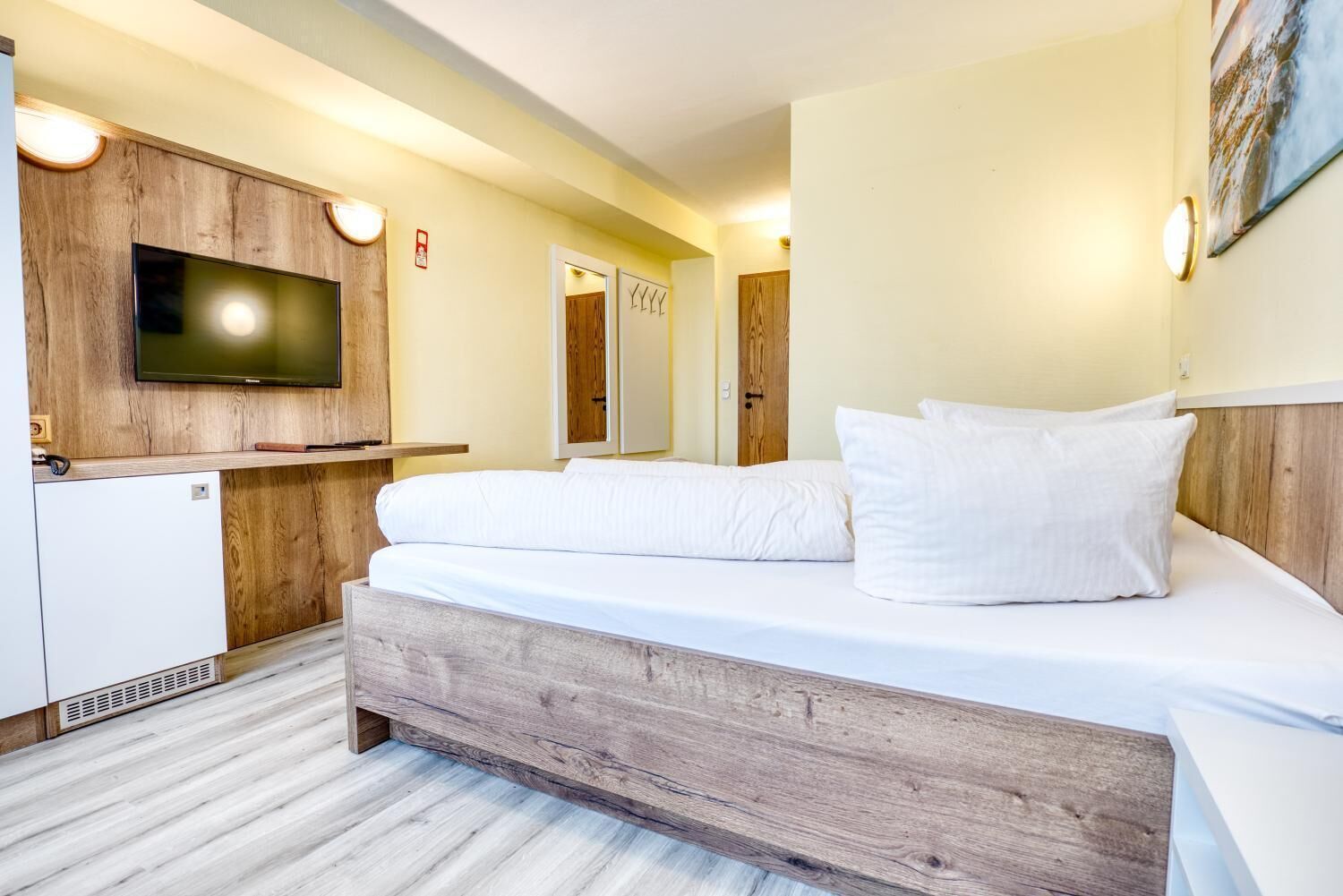 Een moderne slaapkamer met een houten hoofdbord, een flatscreen televisie, een minibar en een groot bed met witte beddengoed.