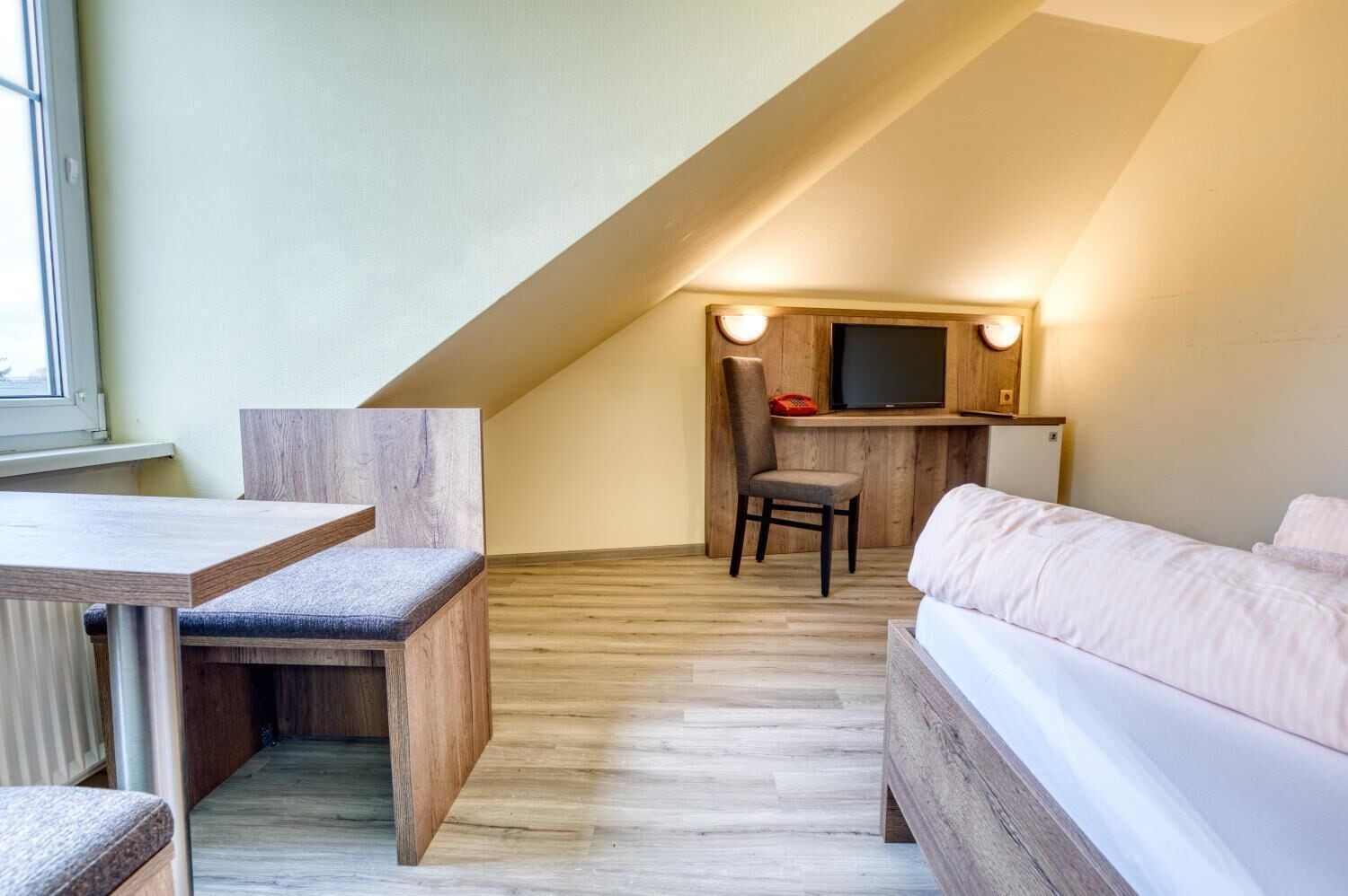 Une chambre confortable située dans les combles, avec un bureau en bois, un fauteuil confortable, un lit et un téléviseur fixé au mur.