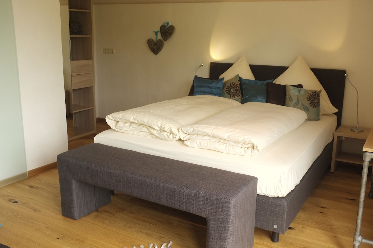 Una cama bien hecha con un banco gris al frente, una mesita de noche con una lámpara y una pared decorada con adornos con forma de corazón.