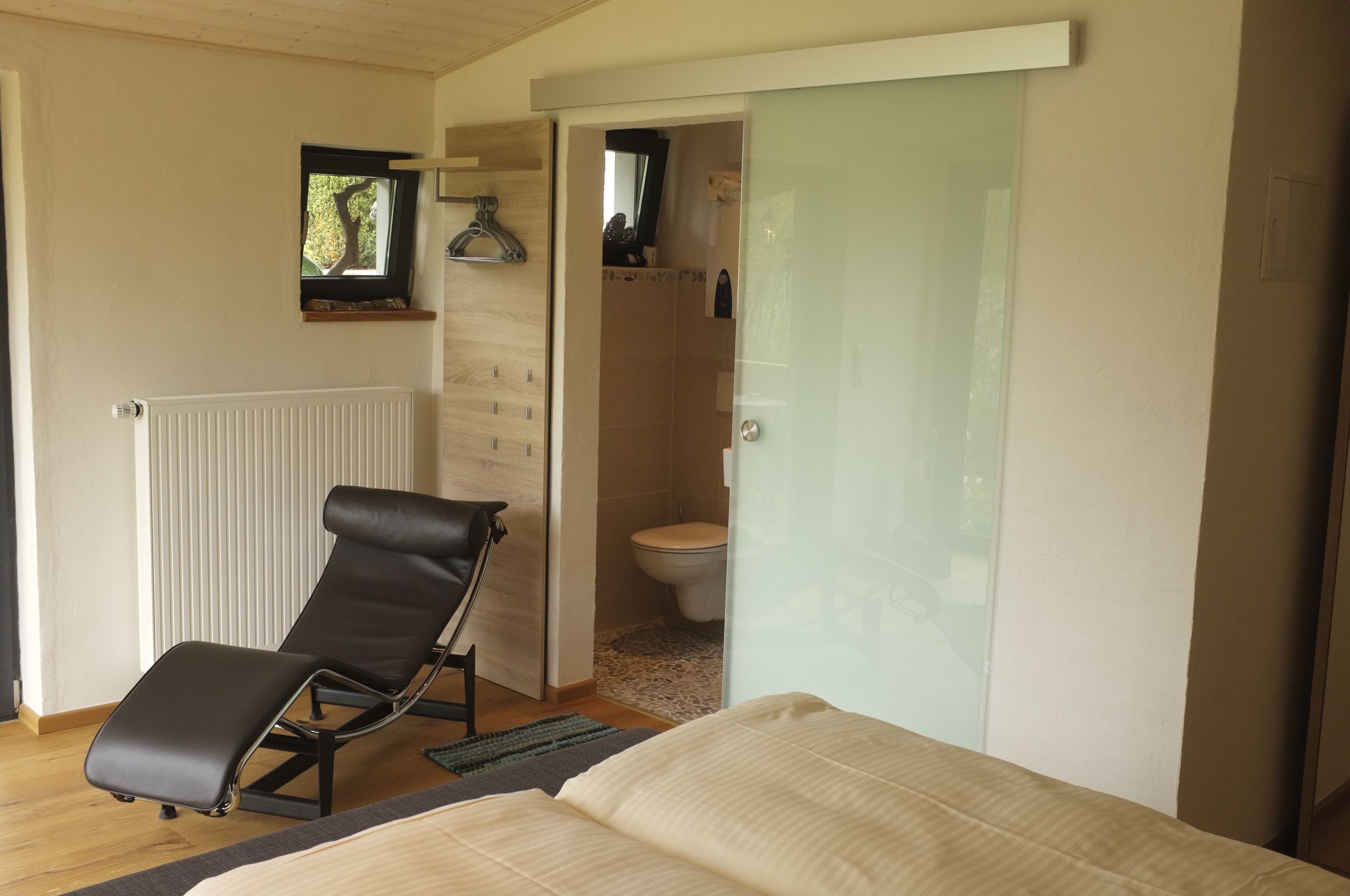 Ein Schlafzimmer mit einem Bett, einem schwarzen Ledersessel, einer Heizung, einem kleinen Fenster und einem durch eine Glastür einsehbaren Badezimmer.
