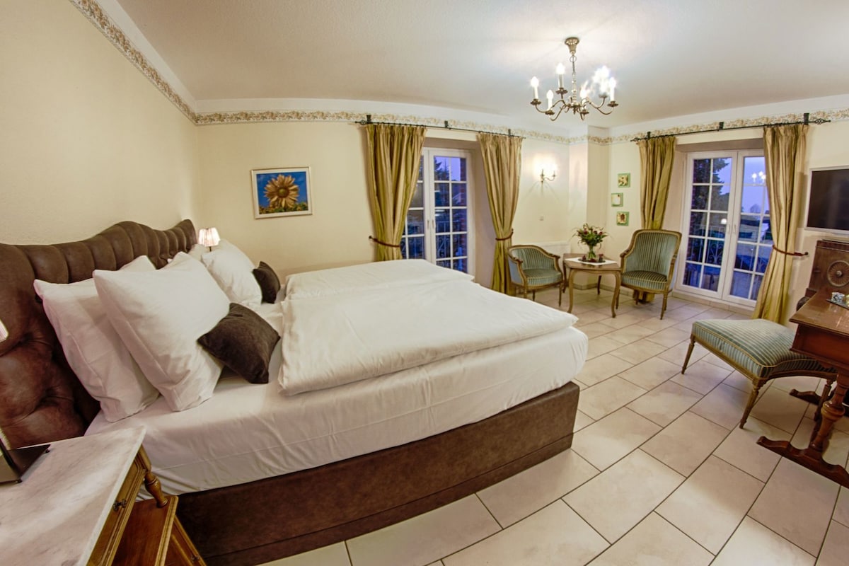 Habitación de hotel con una cama grande, un escritorio, dos sillas, una araña y una ventana con cortinas.