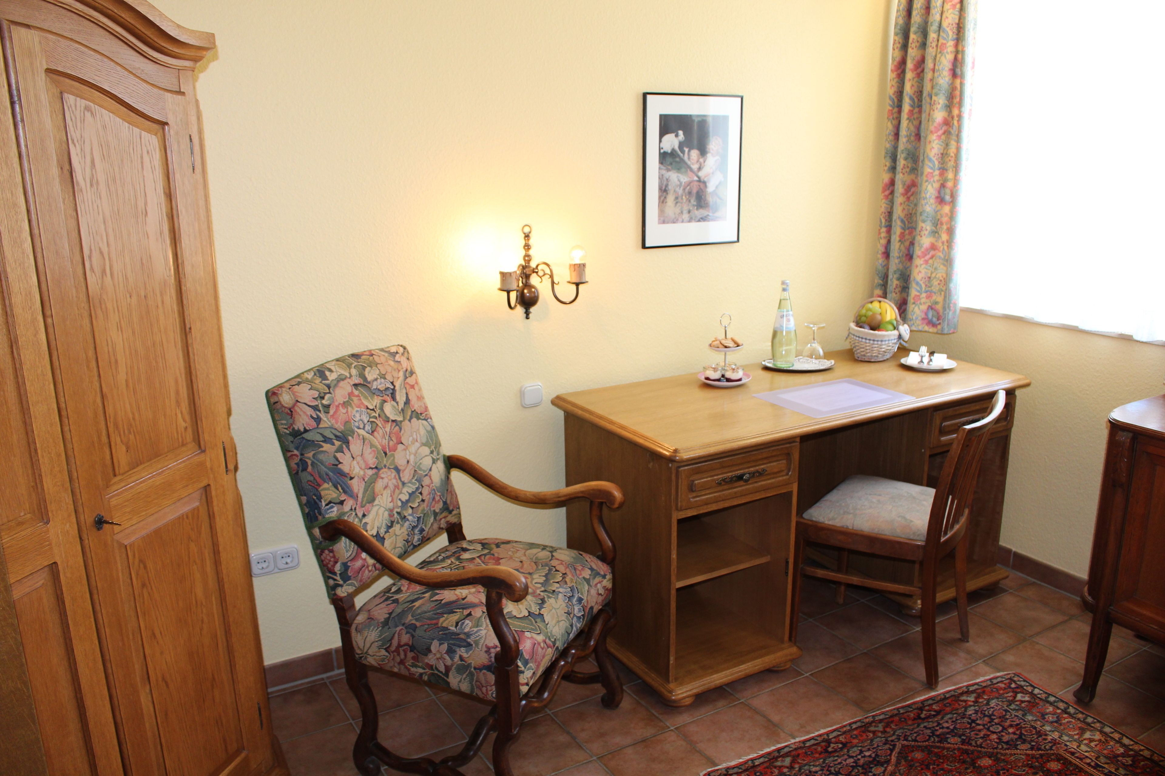 A room with a wooden desk, a floral chair, a wooden wardrobe, a framed picture on the wall, and a window with curtains.