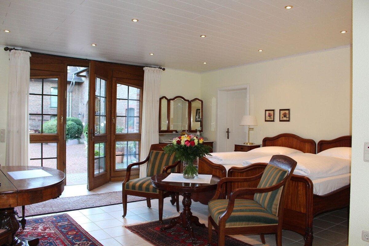 Habitación de hotel con cama, dos sillas, una mesa con un jarrón de flores, escritorio, espejo y vistas al exterior.