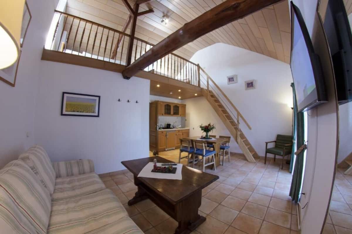 Amplia sala de estar con techo alto, escalera que conduce a un nivel superior, comedor con sillas y mesa, y una cocina al fondo.