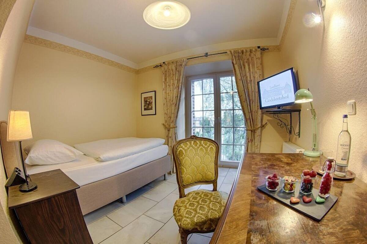Habitación de hotel con cama, una silla, escritorio con lámpara, televisión y ventana con cortinas.
