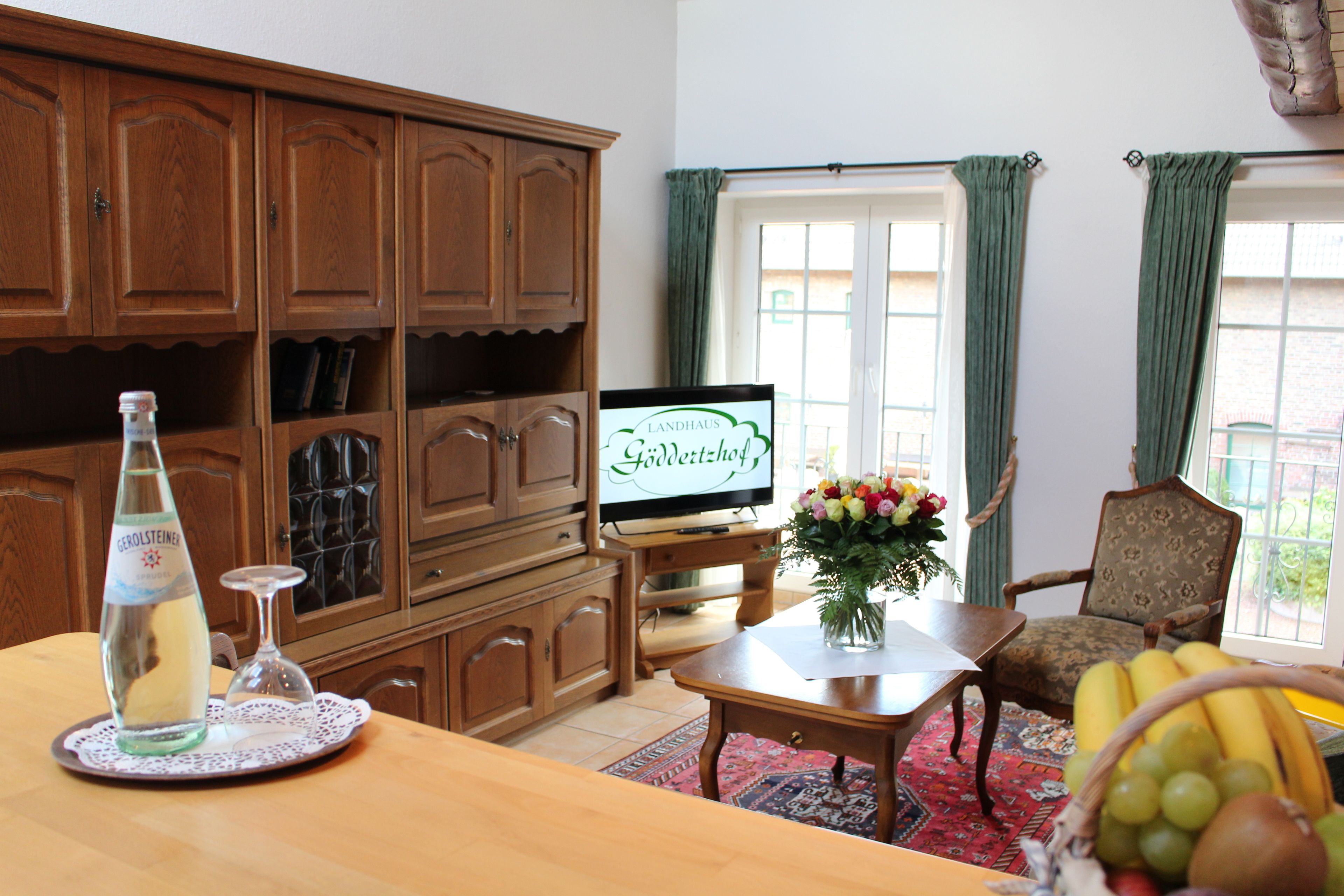 A living room with wooden cabinets, a television, a dining table with a vase of flowers, and a fruit basket.