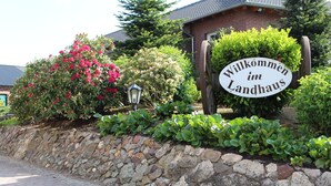 Un muro de piedra con un cartel que dice "Willkommen im Landhaus", rodeado de vegetación exuberante y un edificio de ladrillo.