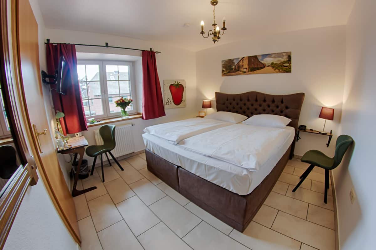 Un dormitorio con una cama grande, dos lámparas de noche, una ventana con cortinas rojas, un radiador y un escritorio con una silla.