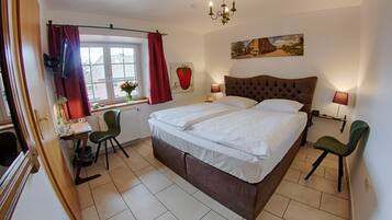 A bedroom with a large bed, two bedside lamps, a window with red curtains, a radiator, and a desk with a chair.