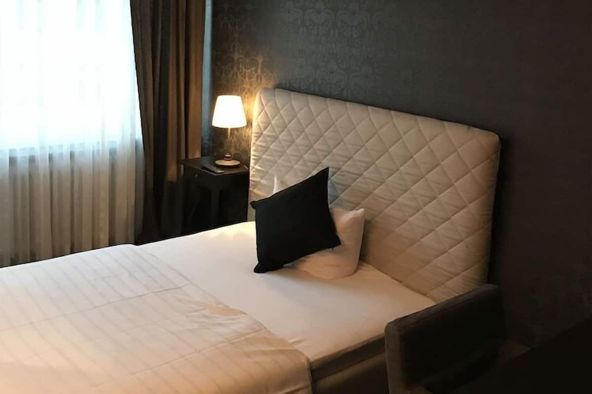 A hotel room with a bed, a nightstand with a lamp, a dark wall, and a window with curtains.