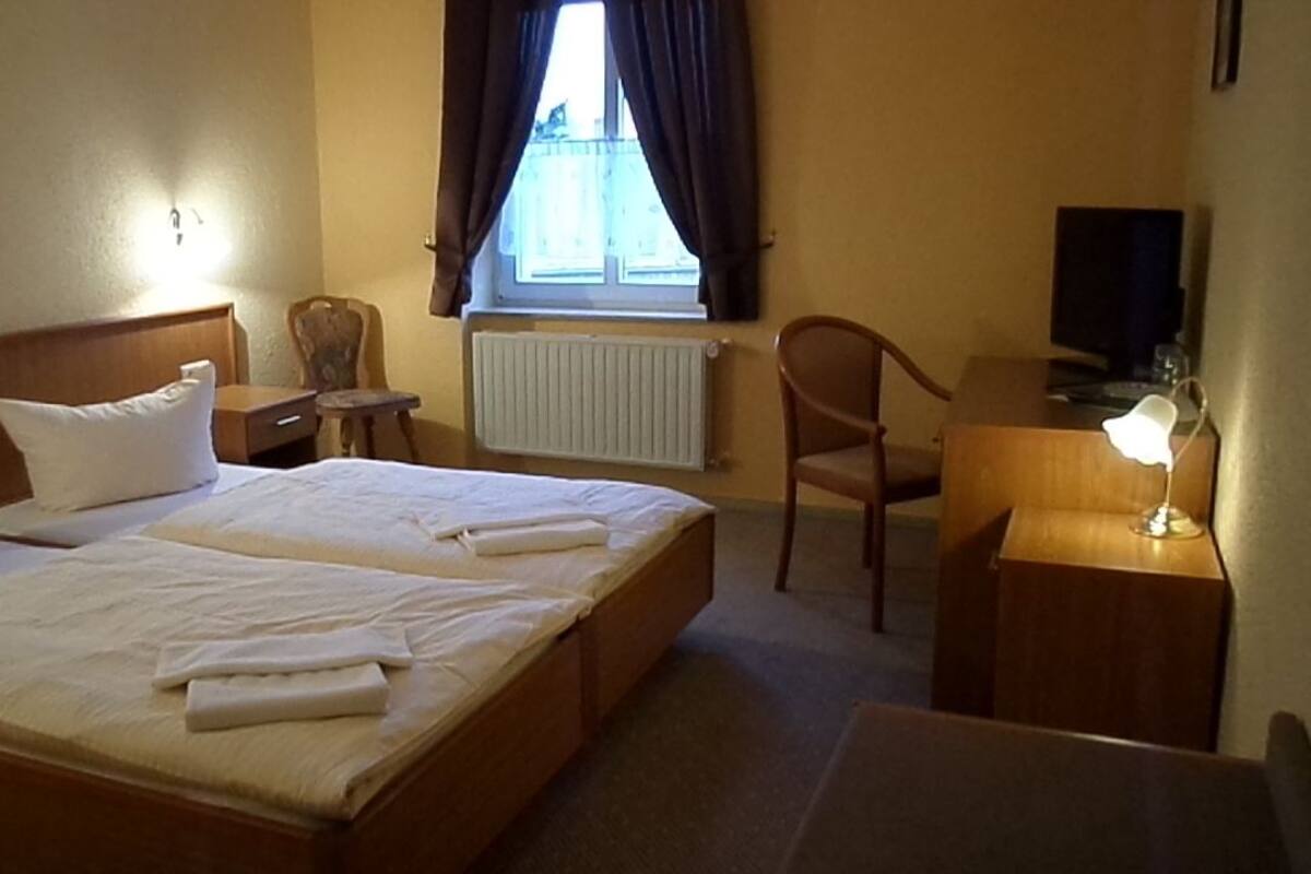 A hotel room with a double bed, a desk with a lamp, a chair, a radiator, and a window with curtains.