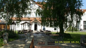 An outdoor seating area with tables and chairs, a building with a tiled roof, and trees in the background. - Hotel Schützenhaus (Bad Dueben)
