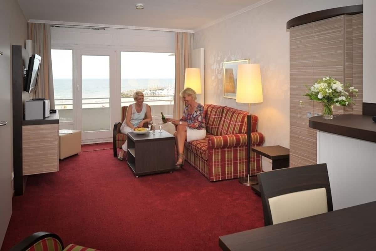 A hotel room with a red carpet, a plaid sofa, a small table with a bottle and glass, a TV on a stand, and a large window with a view of the sea.