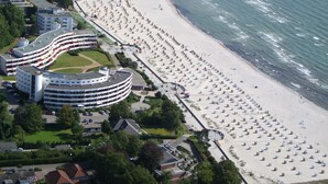 An aerial view of a coastal hotel with a beachfront promenade, multiple-story buildings, and a beach with rows of sun loungers. - Hotel Strandidyll (Groemitz)