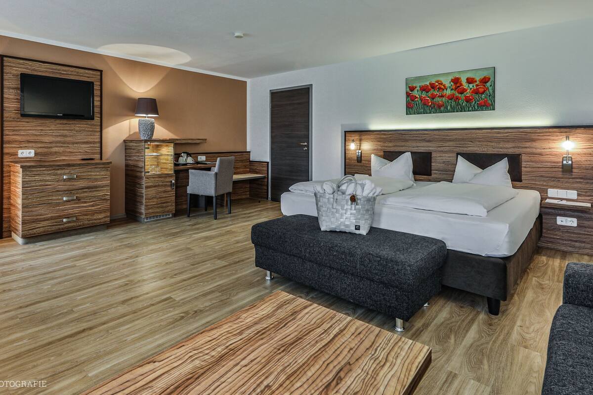 A modern hotel room with a wooden floor, a bed with white linens, a desk with a chair, a TV mounted on the wall, and a painting of red flowers.