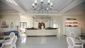 A hotel lobby with a reception desk, a chandelier, a blue sofa, and white chairs. - Olympic Kosma Hotel (Kassandra)