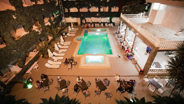 An indoor pool area with multiple lounge chairs and tables, surrounded by greenery and a patterned floor design. - Olympic Kosma Hotel (Kassandra)