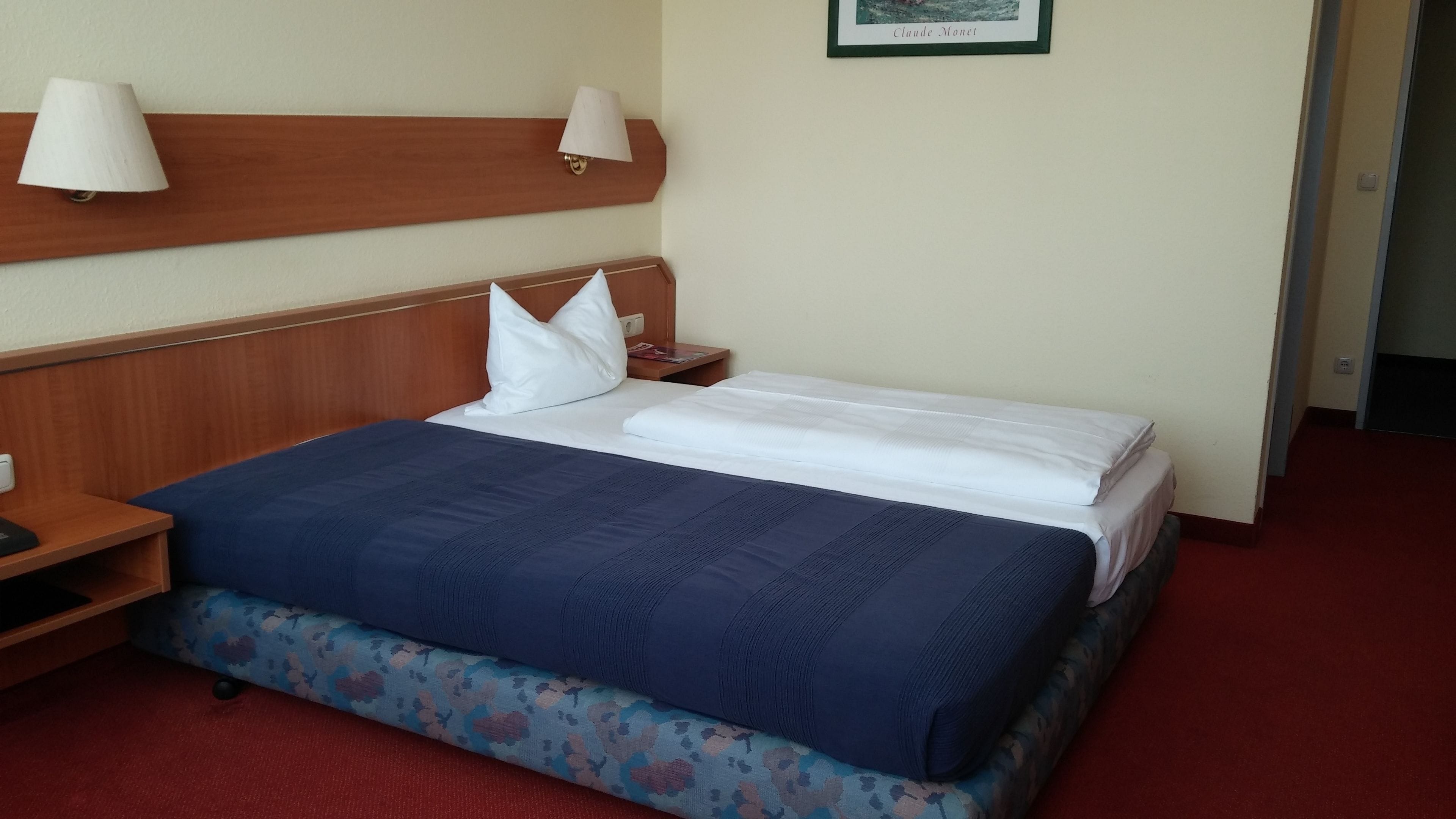 A hotel room with a neatly made bed, a wooden headboard, a nightstand, and a wall-mounted lamp.