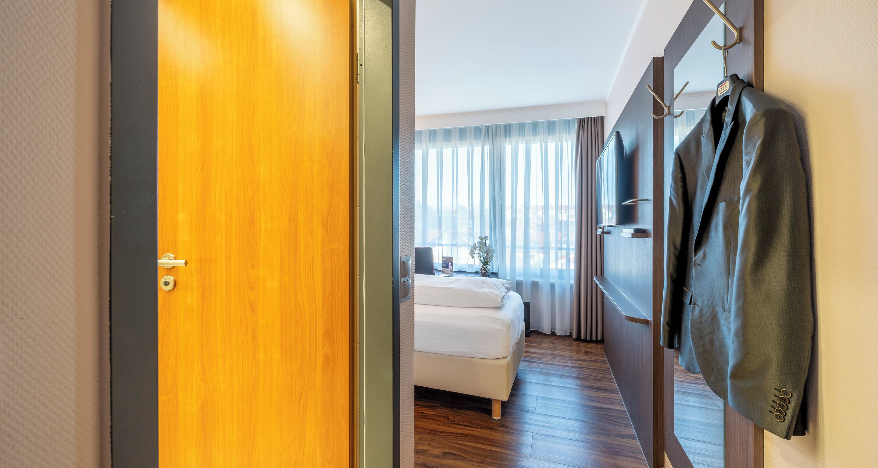 Habitación de hotel con puerta de madera, cama, televisor y perchero con una chaqueta.