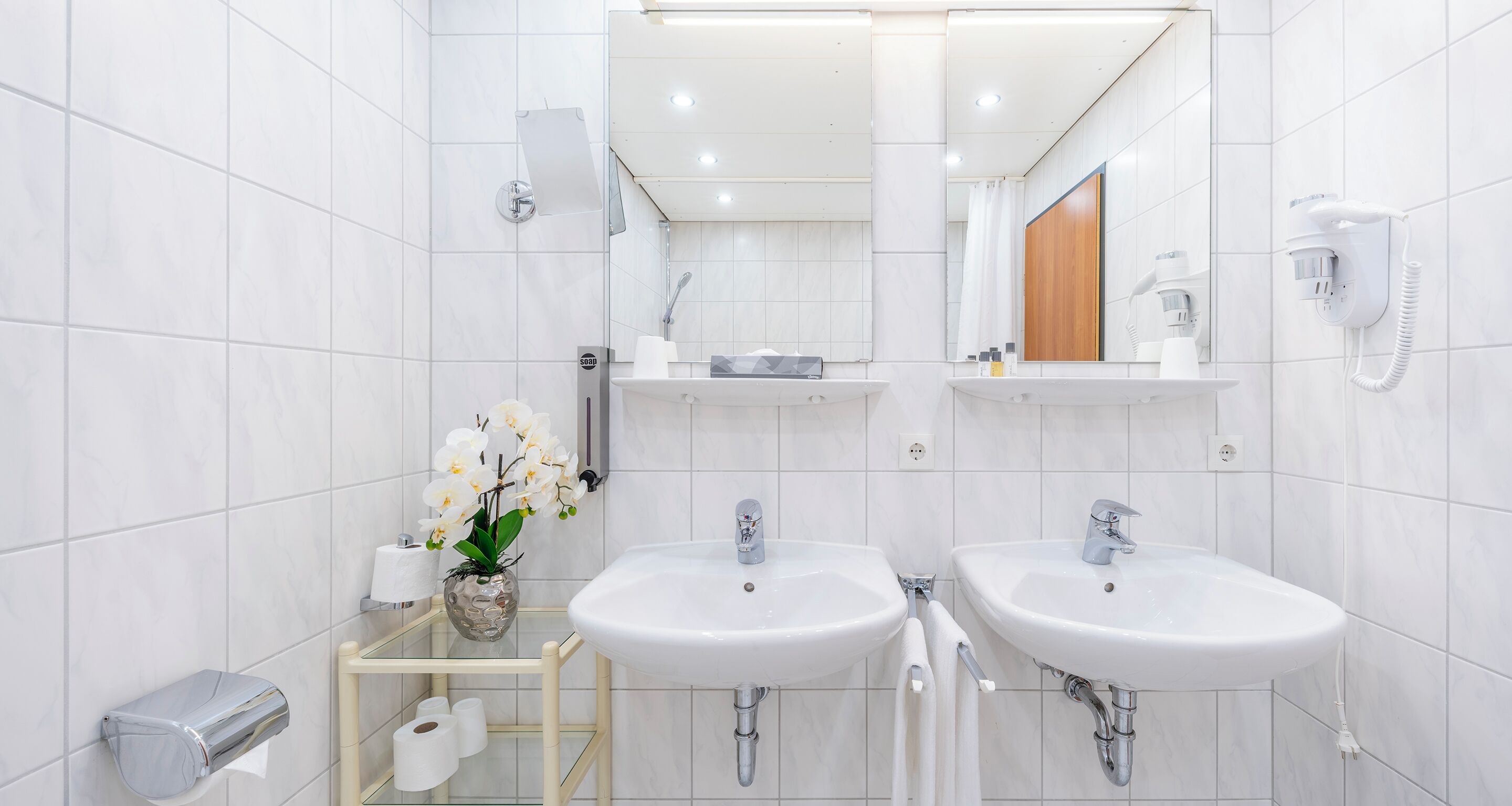 Un baño moderno con dos lavamanos, un espejo, toallero, dispensador de papel higiénico y secador de pelo.