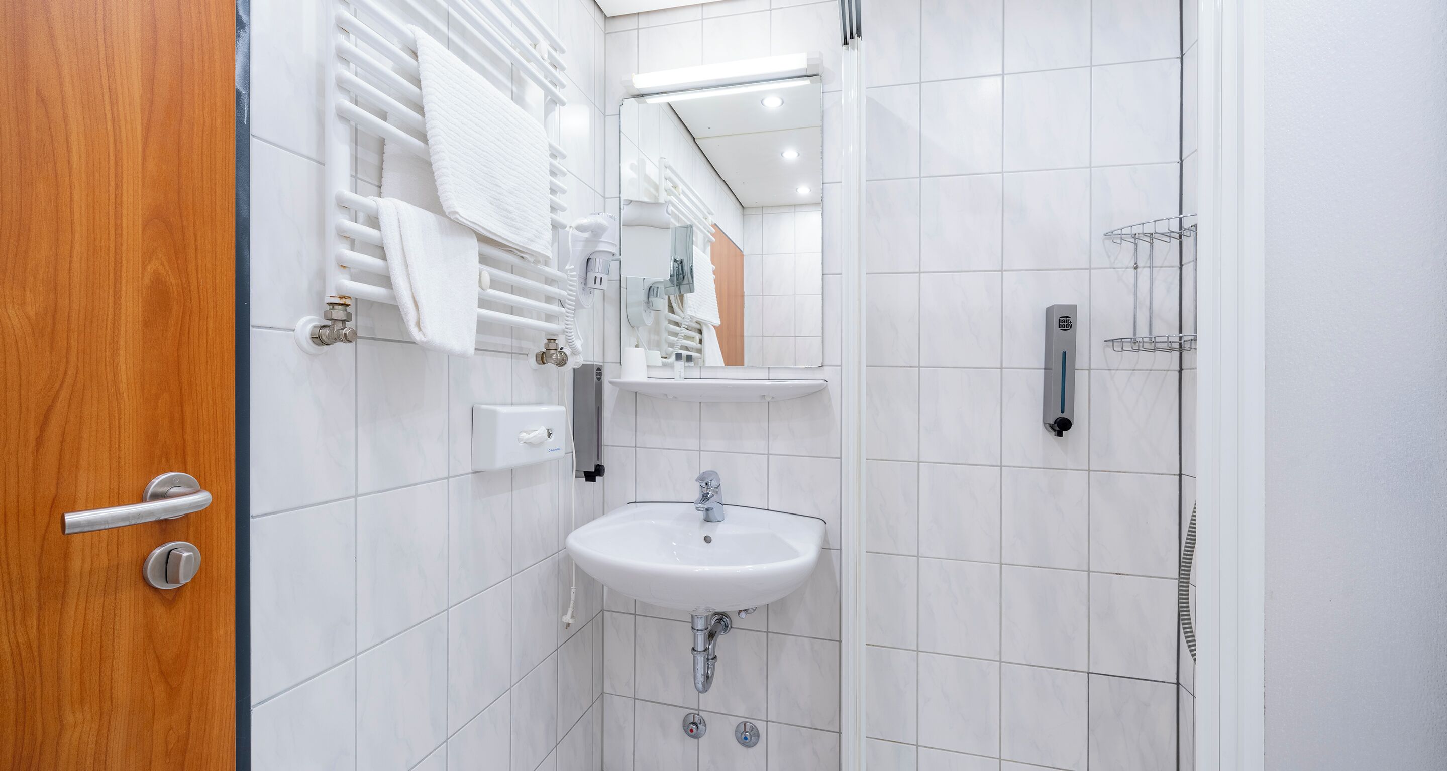 Ein Badezimmer mit Holztür, weißen Fliesenwänden, einem Waschbecken, einem Spiegel und einem Handtuchhalter.