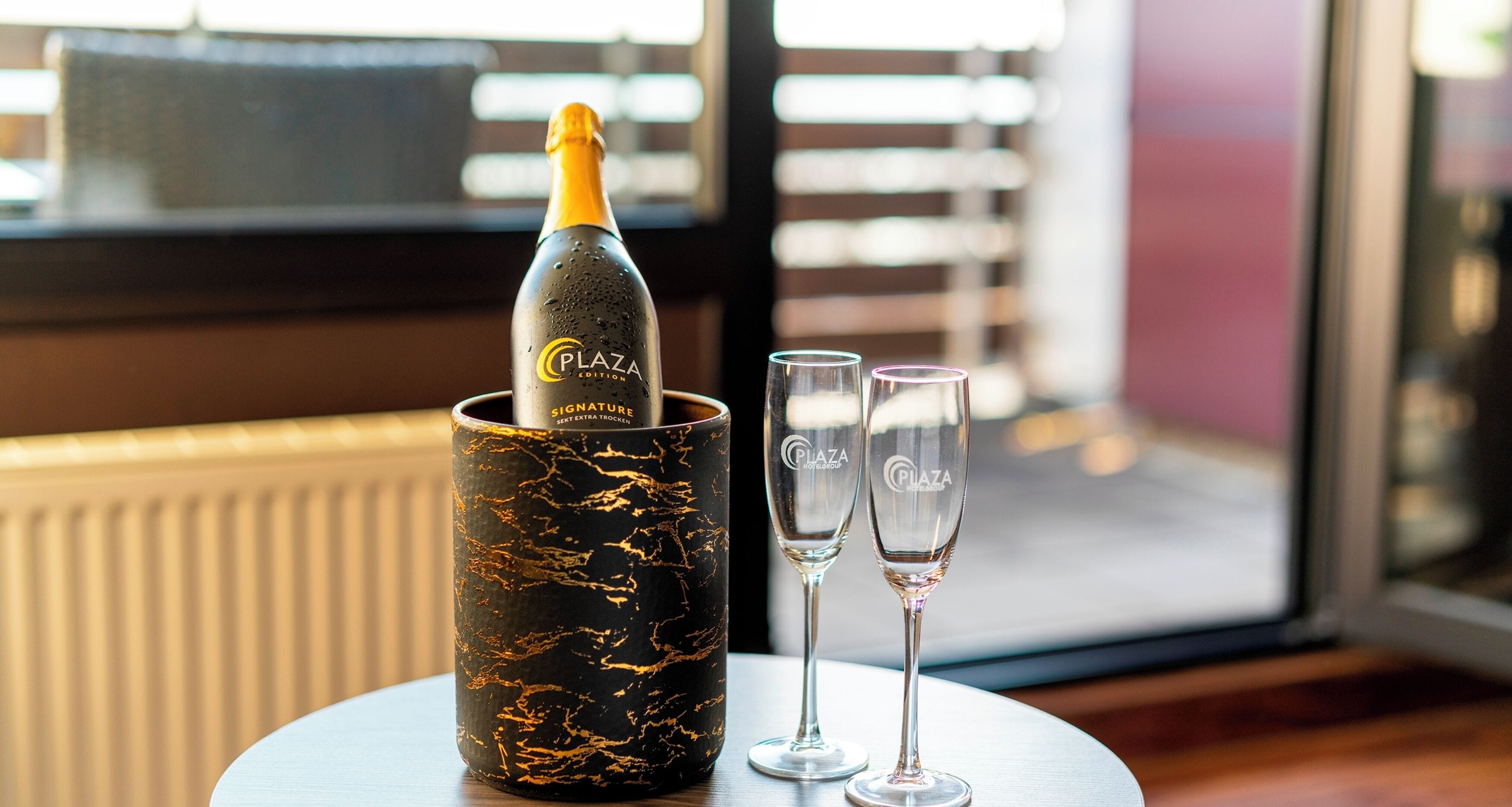 A bottle of Plaza Signature Champagne in a black marble-patterned container, two empty champagne flutes, and a window with blinds in the background.
