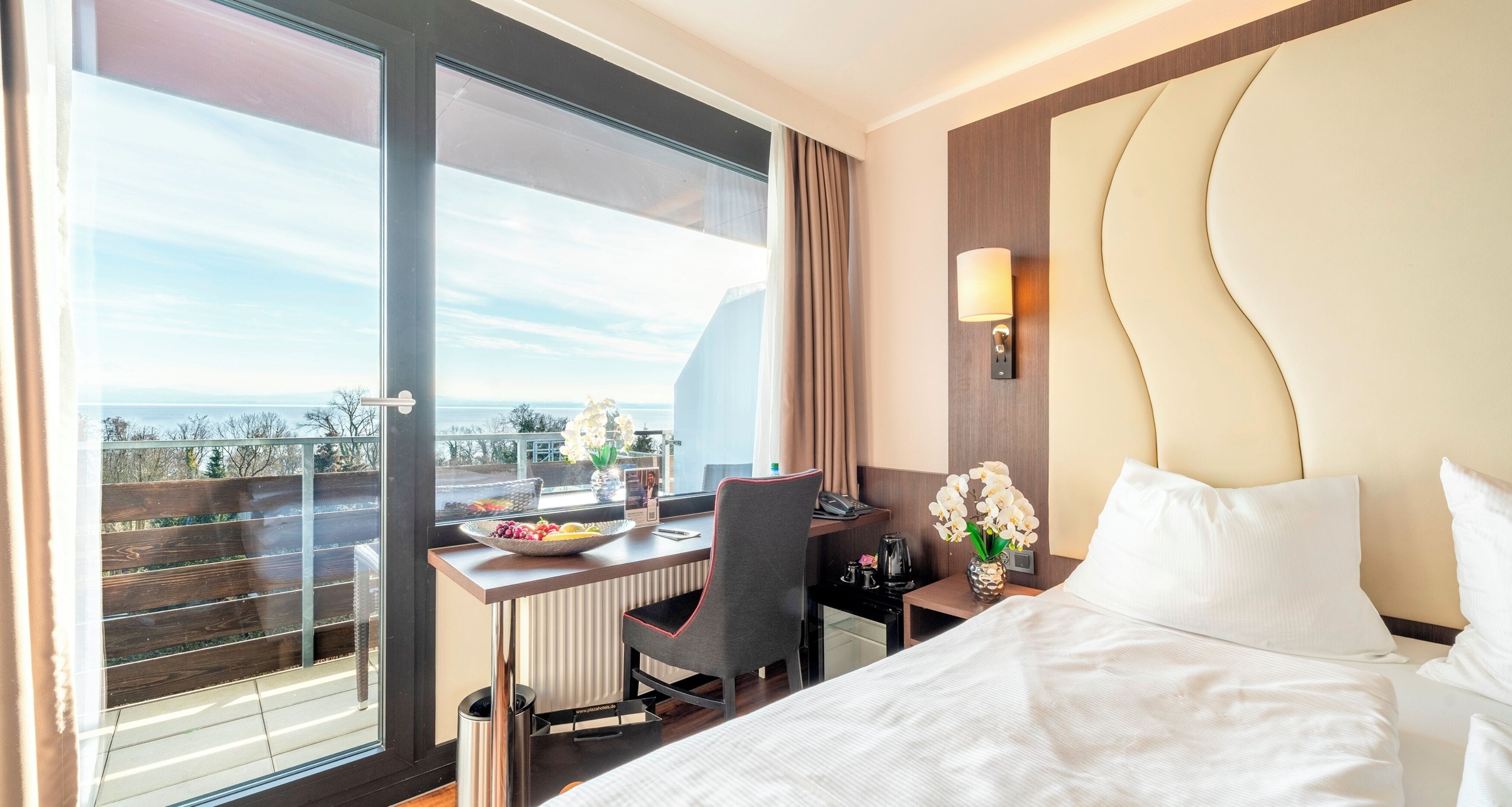 A hotel room with a large window, a desk with a bowl of fruit, a chair, a bed with white linens, and a vase of flowers.