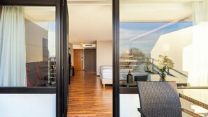 Ein Hotelzimmer mit Holzboden, einer Couch, einem Esstisch mit Stühlen, einer kleinen Küchenzeile und einem großen Fenster mit Vorhängen.
