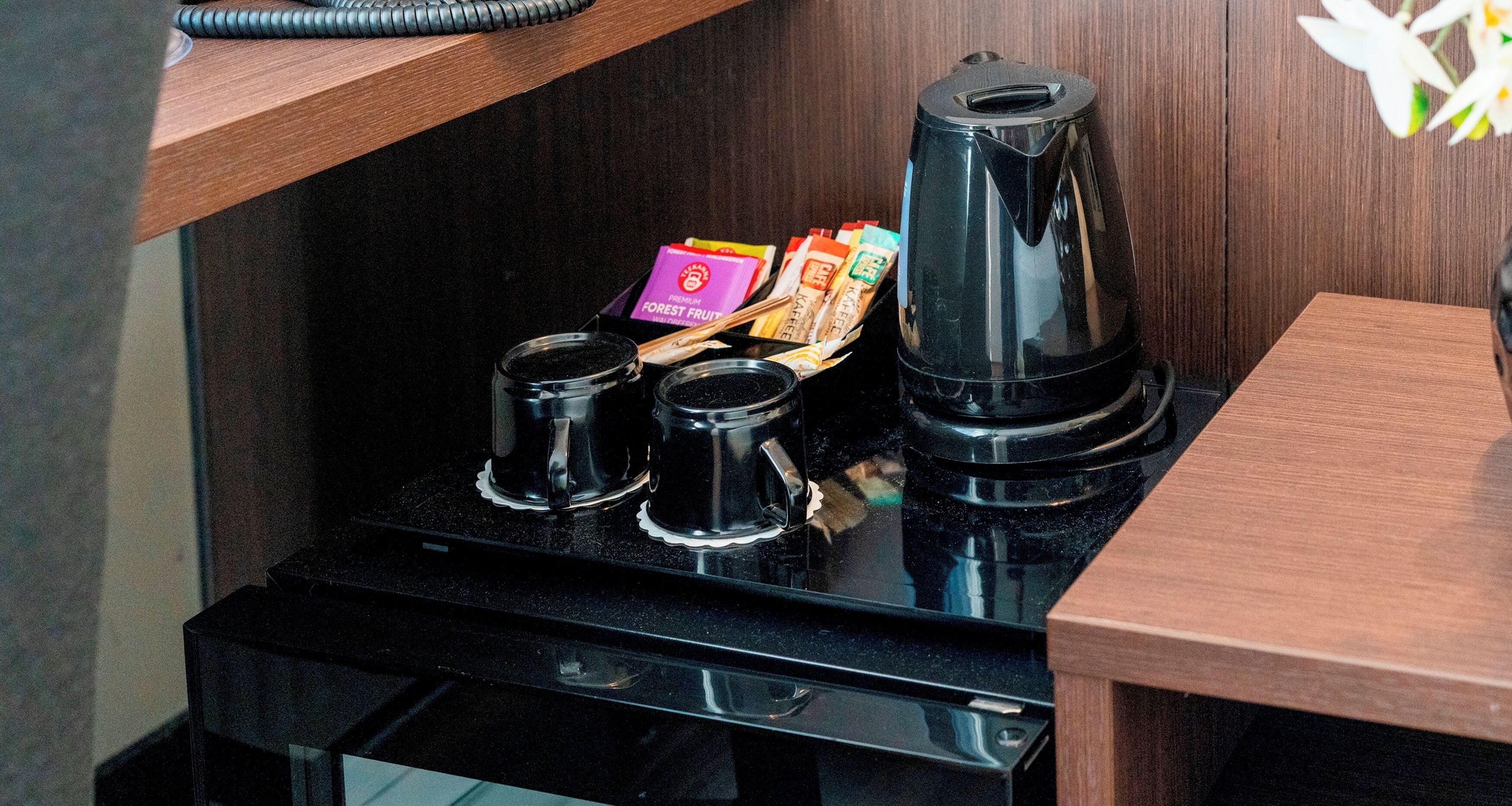 A compact kitchen area with a coffee maker, two small cups, and a box of coffee pods on a black countertop.