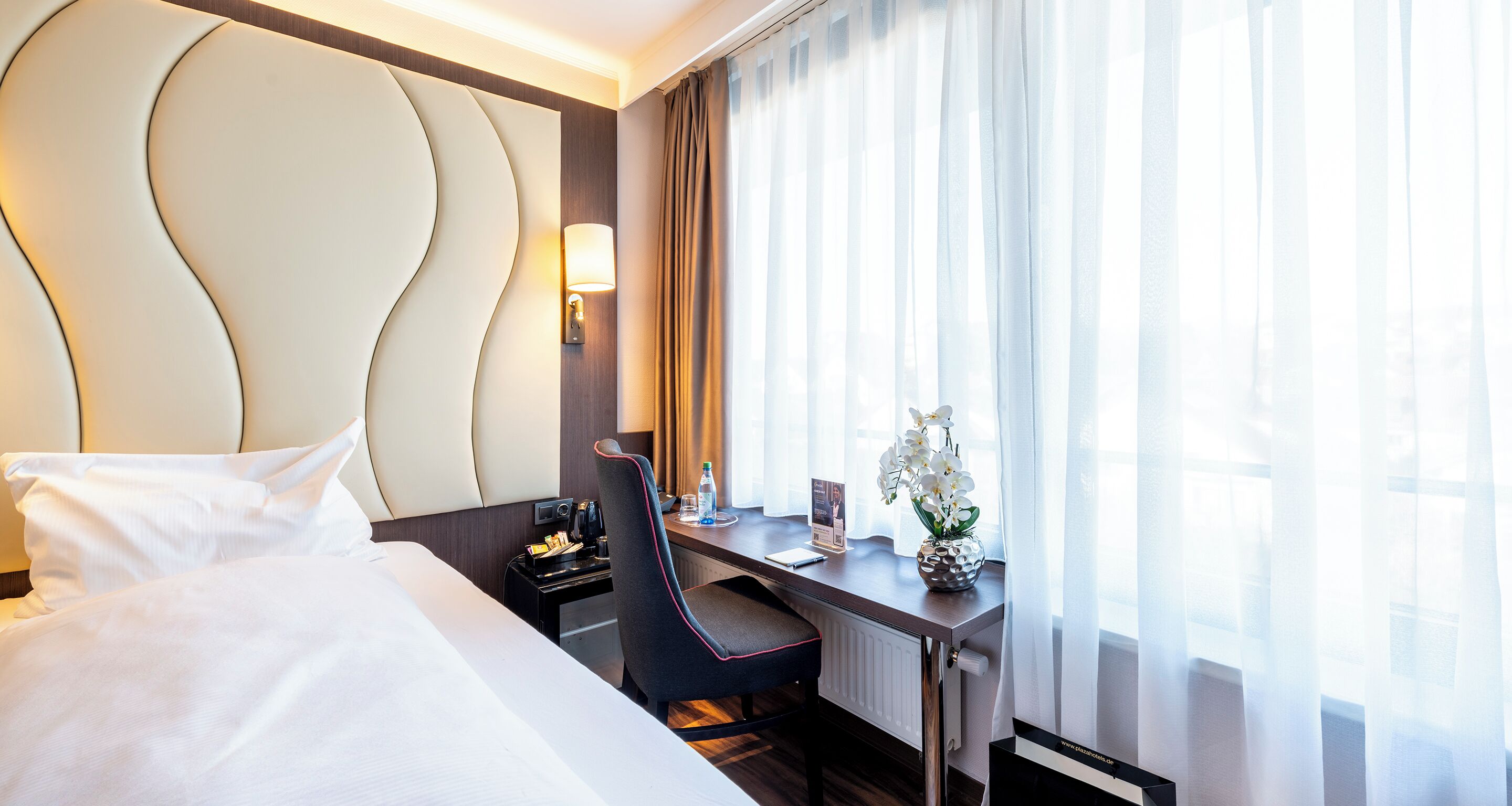 Una habitación de hotel con cama, escritorio con silla, lámpara, florero con flores y ventana con cortinas.