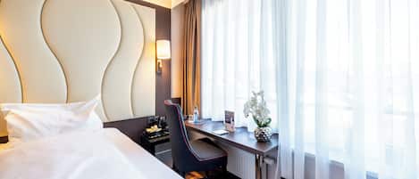 Una habitación de hotel con cama, escritorio con silla, lámpara, florero con flores y ventana con cortinas.