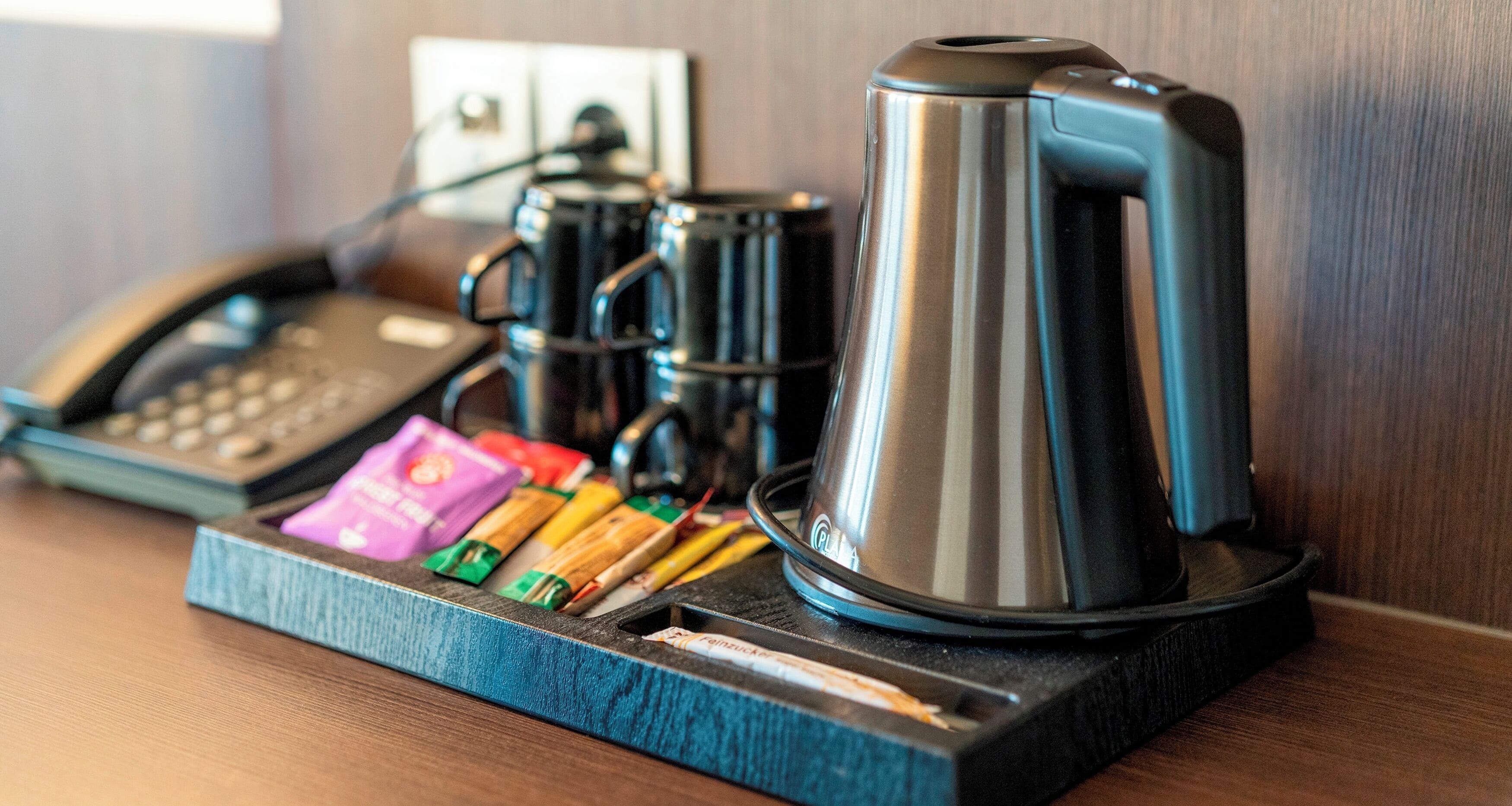 A coffee station with a coffee maker, a phone, and some snacks on a wooden surface.