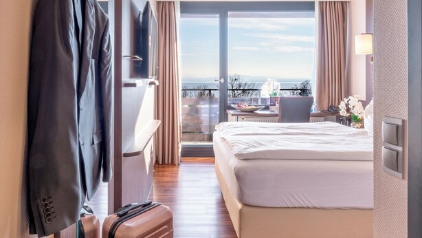 Ein Hotelzimmer mit Bett, Schreibtisch mit Stuhl, Koffer und Meerblick durch eine Glasdoor.