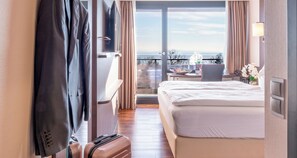 Een hotelkamer met een bed, een bureau met stoel, een koffer en uitzicht op zee door een glazen deur.
