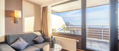 Ett modernt hotellrum med en grå soffa, ett litet runt bord med en flaska champagne och två glas, ett stort fönster med utsikt över staden och en vägglampa.