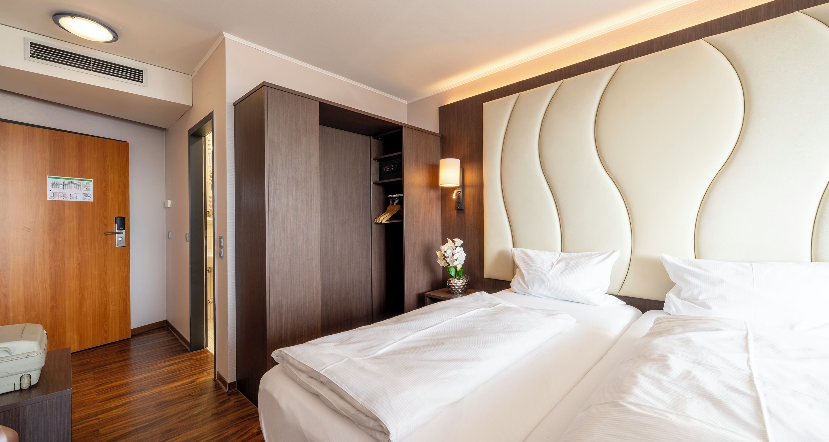 A hotel room with a large bed, a wardrobe, a bedside table with a lamp, and a wooden door.