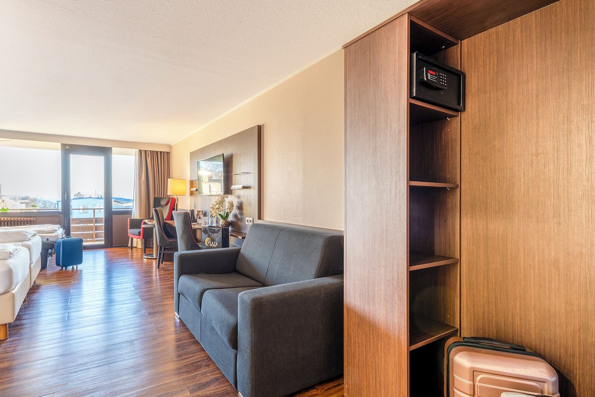 Une chambre d’hôtel avec un plancher en bois, un canapé gris, un coin repas avec une table et des chaises, et une vue dégagée sur l’extérieur grâce à une grande fenêtre.