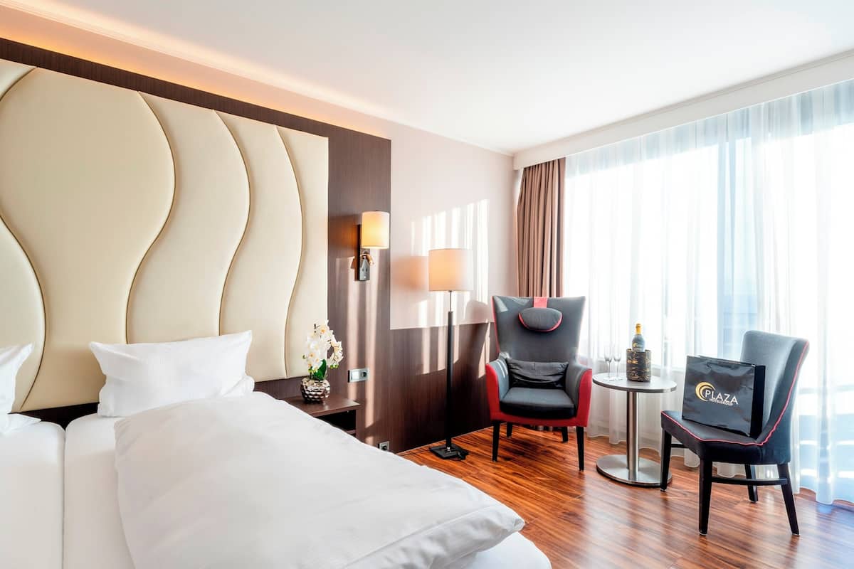 Une chambre d’hôtel avec un grand lit, deux fauteuils, une petite table avec une bouteille et des verres, et une vue sur la ville grâce à de grandes fenêtres.