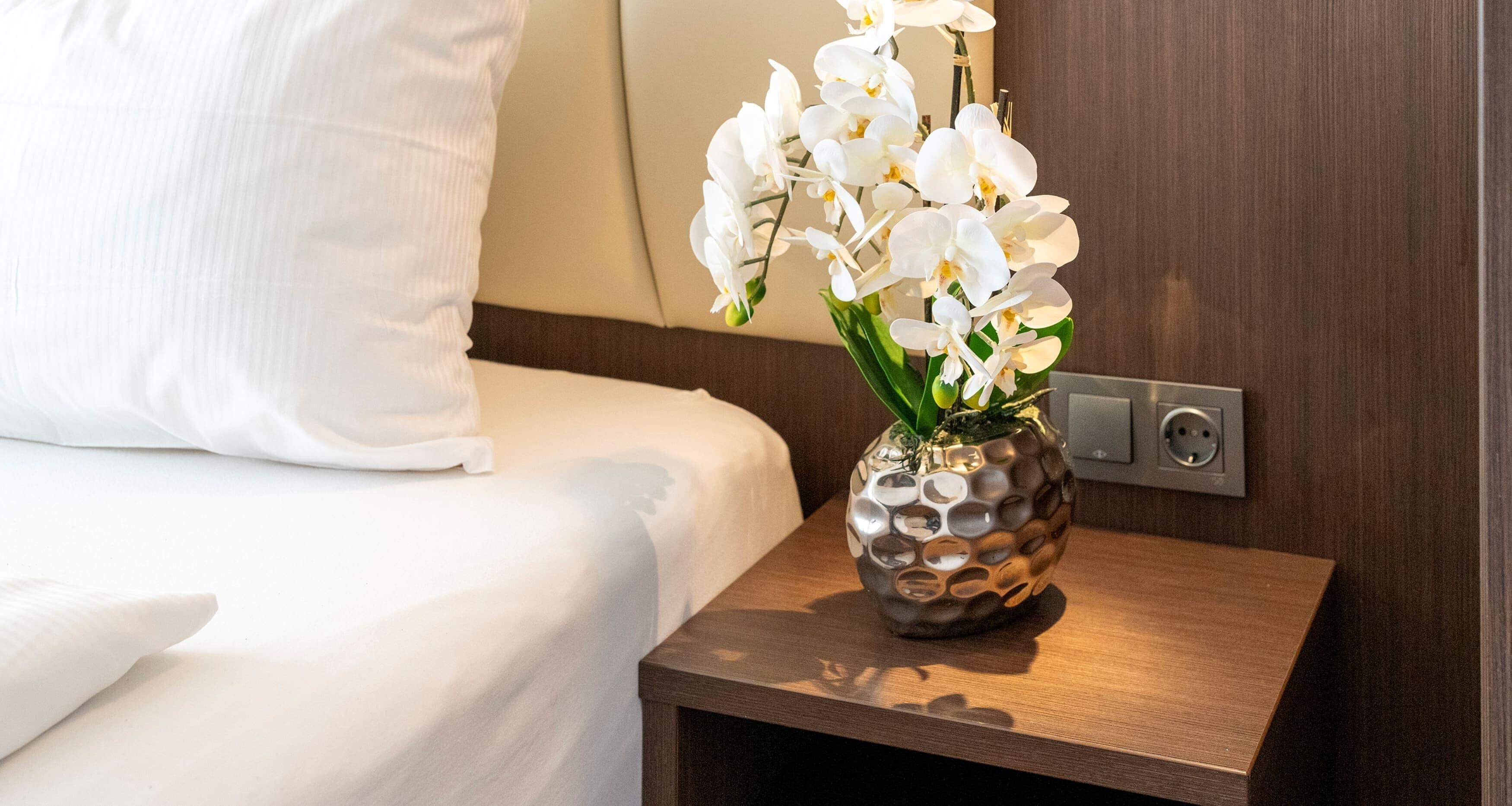 Habitación de hotel con una cama bien hecha, una mesita de noche de madera con un jarrón decorativo de orquídeas blancas y un enchufe de pared.