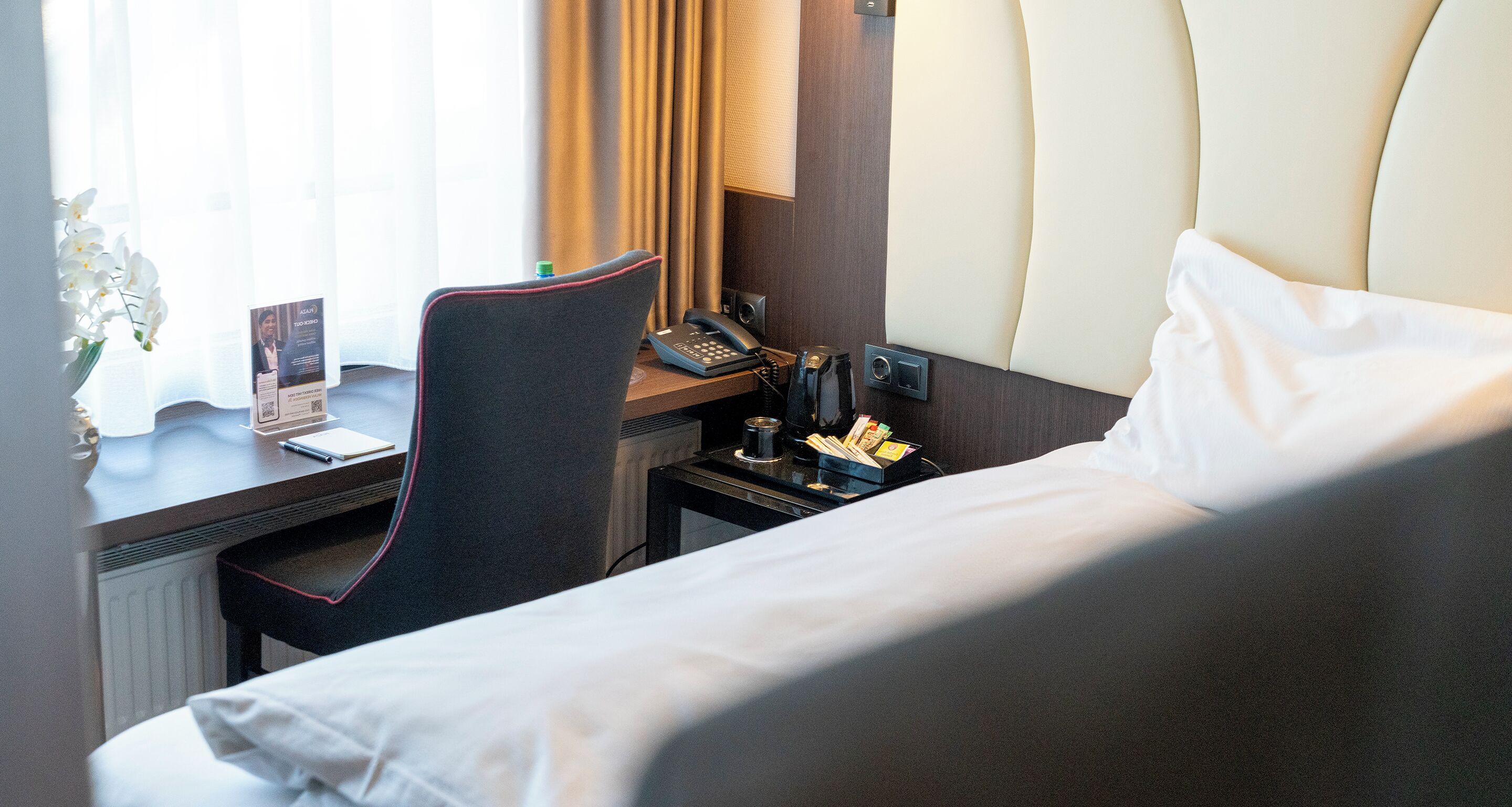 Hotellihuone, jossa on sänky, kirjoituspöytä, jossa on puhelin, tuoli, pieni pöytä, jossa on lamppu, ja ikkuna, jossa on verhot.